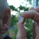 A hand holds a small hearing aid beside an ear in a historical close-up photograph